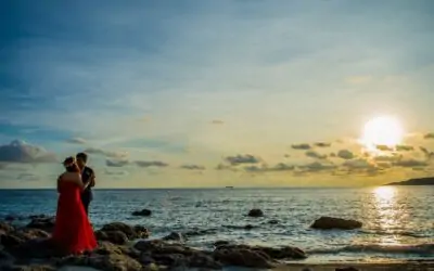 A couple embracing on a rocky beach at sunset, with the ocean in the background.