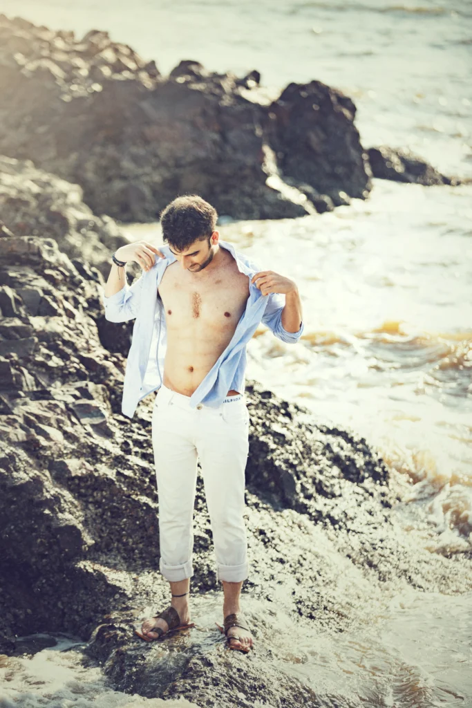 A person standing on rocky terrain near the water’s edge, holding open a light blue shirt, with waves crashing against the rocks in the background.