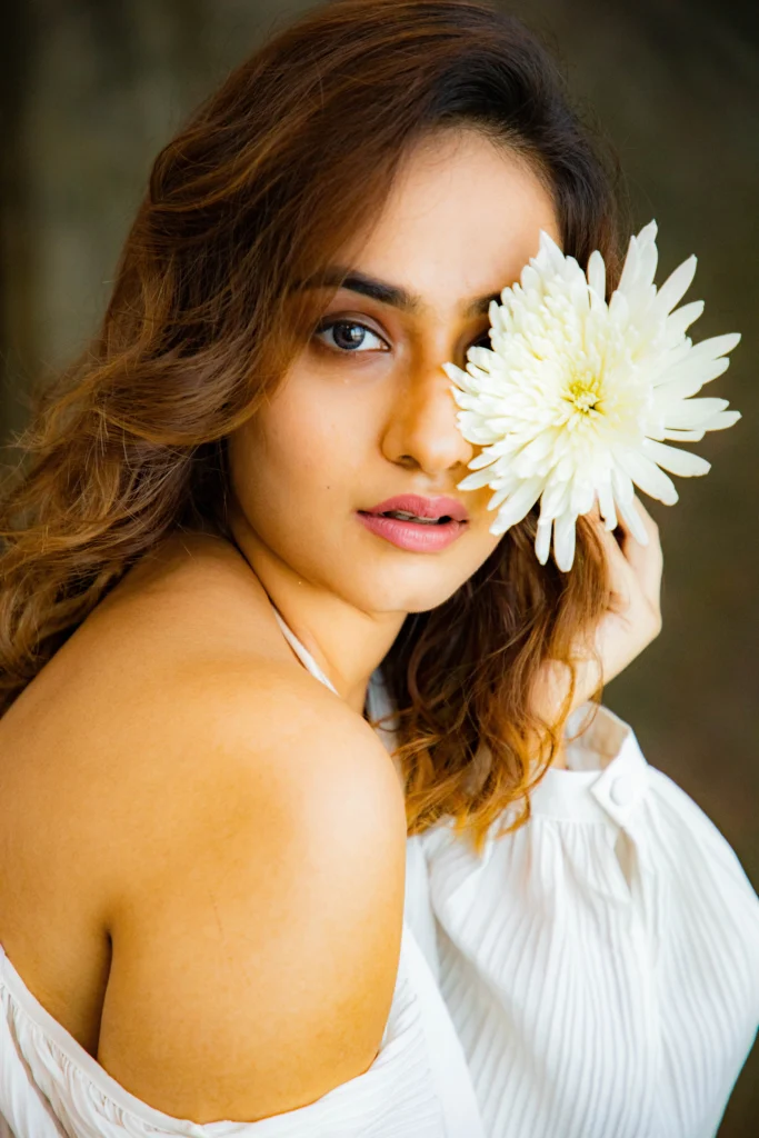 A person holding a large white flower to their face, which is obscured by the flower, wearing a white top against a blurred natural background.