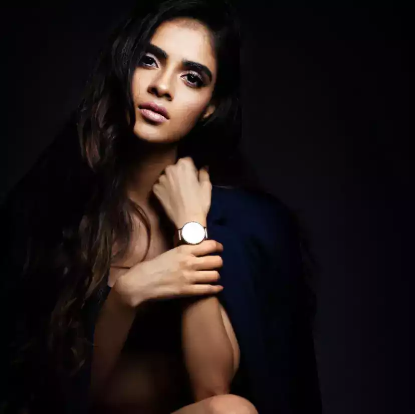 A woman with long dark hair poses elegantly, wearing a watch and a dark blue garment, against a black background.