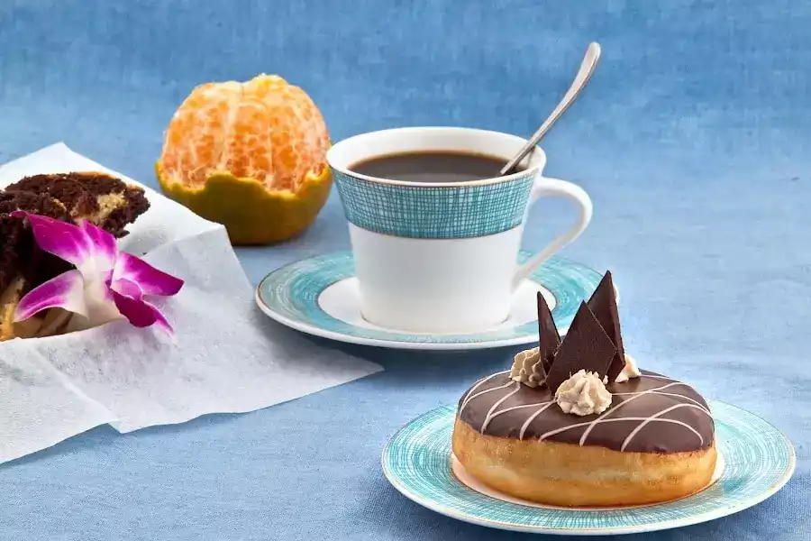 A beautifully arranged breakfast scene featuring a chocolate donut, a slice of cake, a cup of coffee, and a peeled orange.