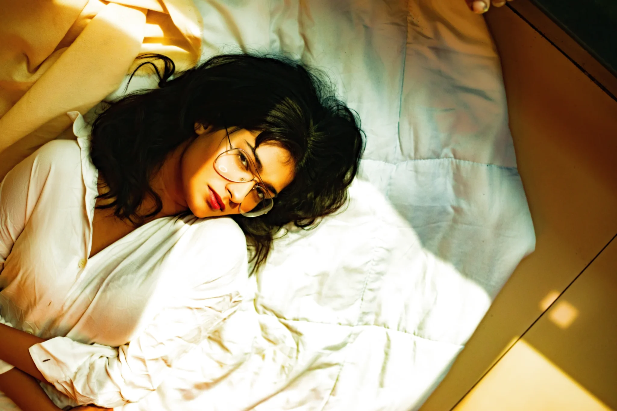 A woman with glasses lying on a bed, looking thoughtfully at the camera.