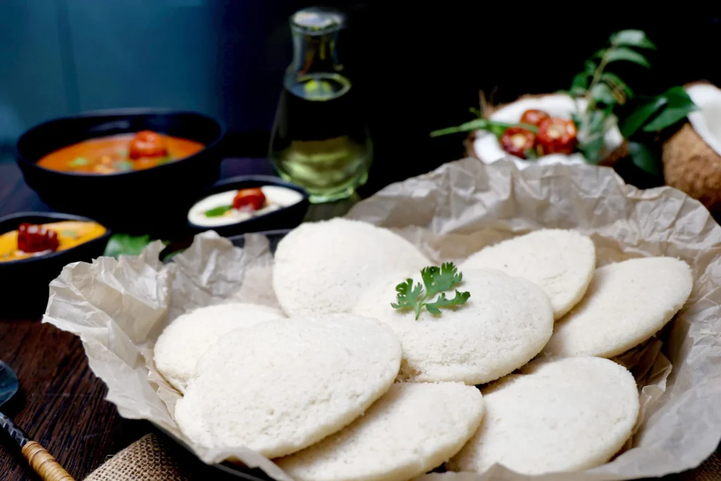 A close-up image of five white, round idlis garnished with a green leaf, placed on a brown paper inside a black dish.