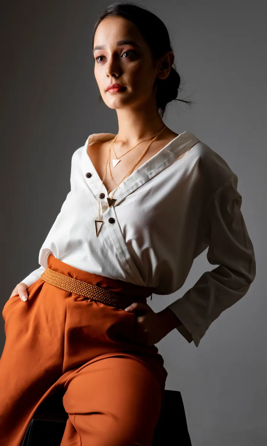 A person standing with one hand on hip, wearing a white shirt and brown colored trousers against a neutral background