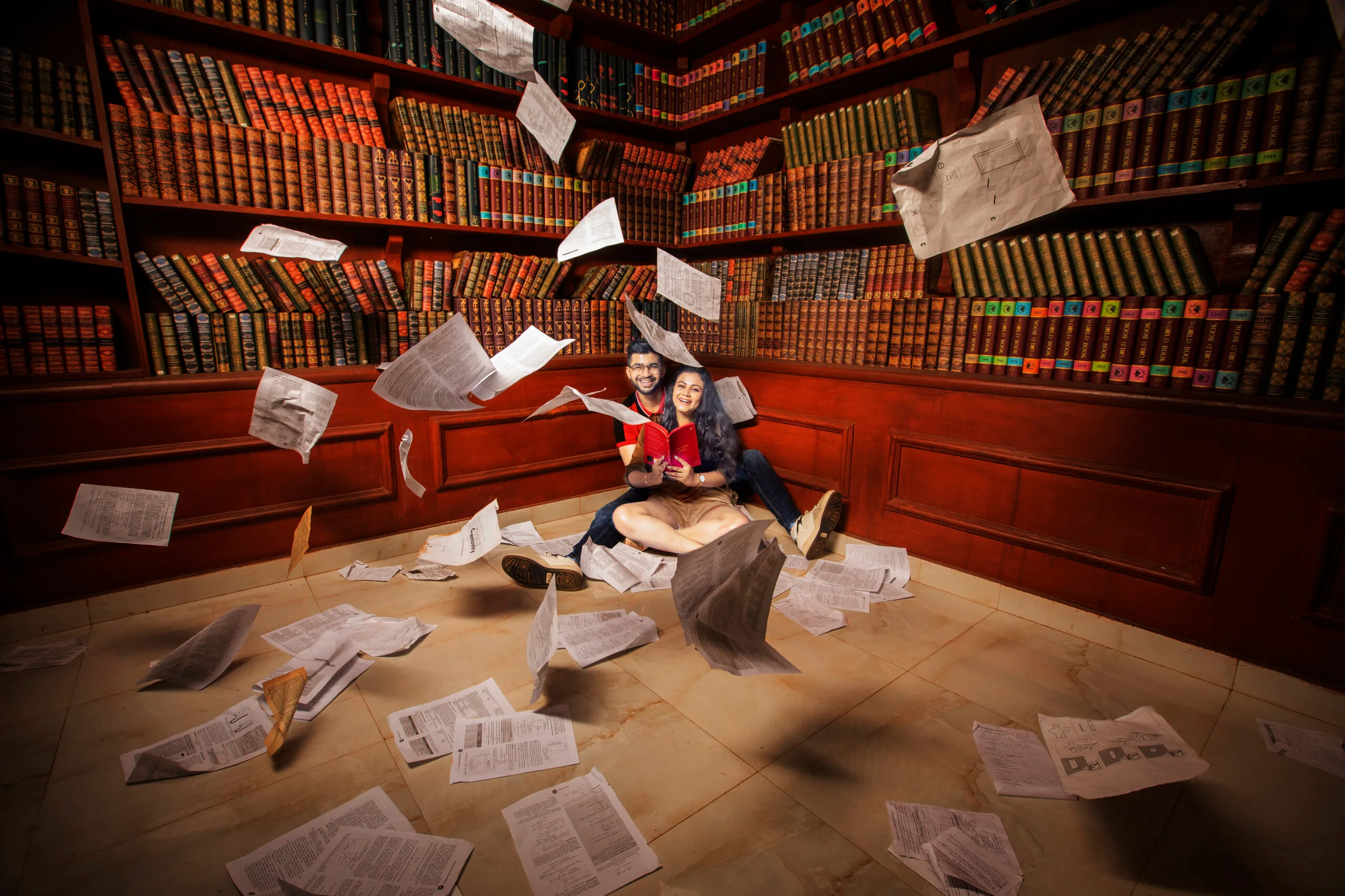A creative pre-wedding photography shoot by students at MDPS, featuring a couple sitting on the floor surrounded by rows of classic bookshelves with flying sheets of paper.