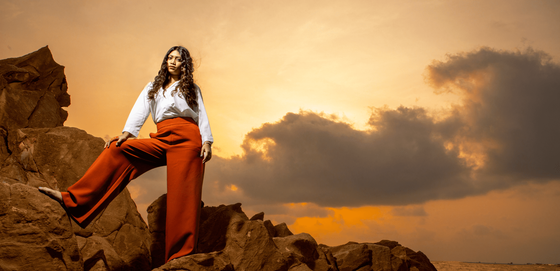 Woman in a white blouse and brown wide-legged pants standing on rocks at sunset, with a colorful sky and clouds in the background.