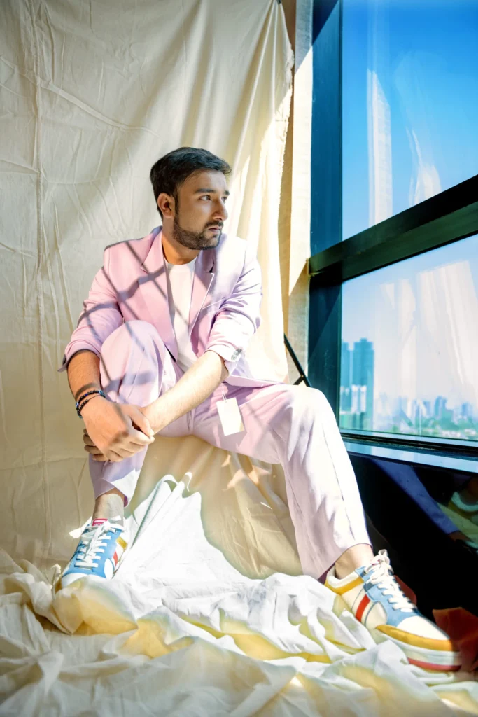 A person in a pink suit sitting with crossed legs in front of a large window with a cityscape view, creating an artistic and modern vibe for the gallery section.