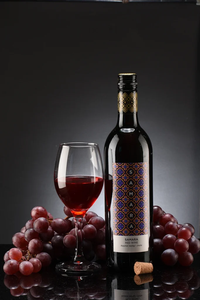 A bottle of red wine with a decorative label next to a filled wine glass, surrounded by clusters of red grapes on a reflective surface with a dark background.