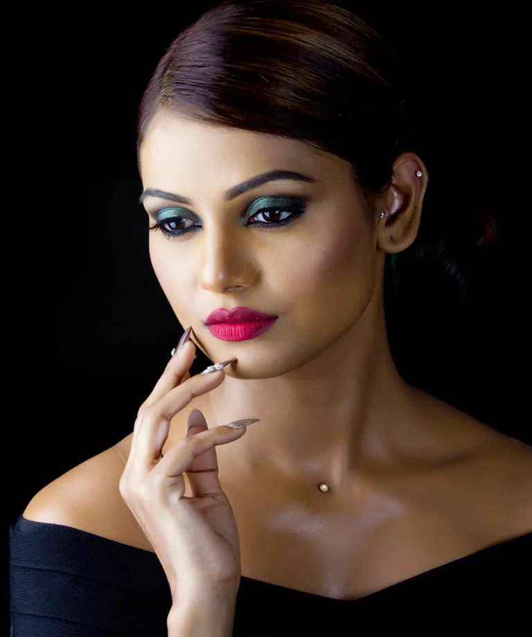 Close-up portrait of a woman with bold red lips and green eye makeup, touching her chin against a black background.