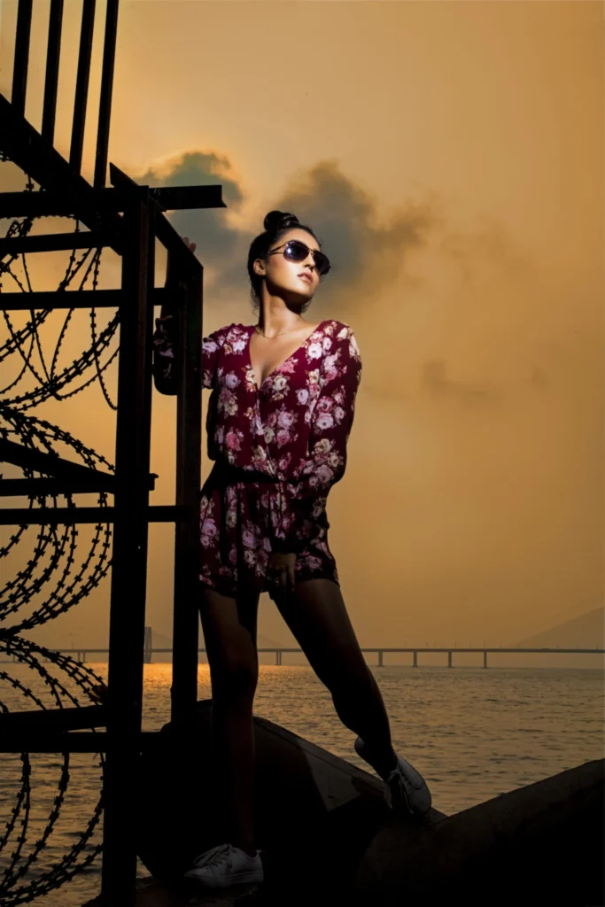 A girl stands against a vibrant sunset, leaning on a metal structure with barbed wire by the sea.