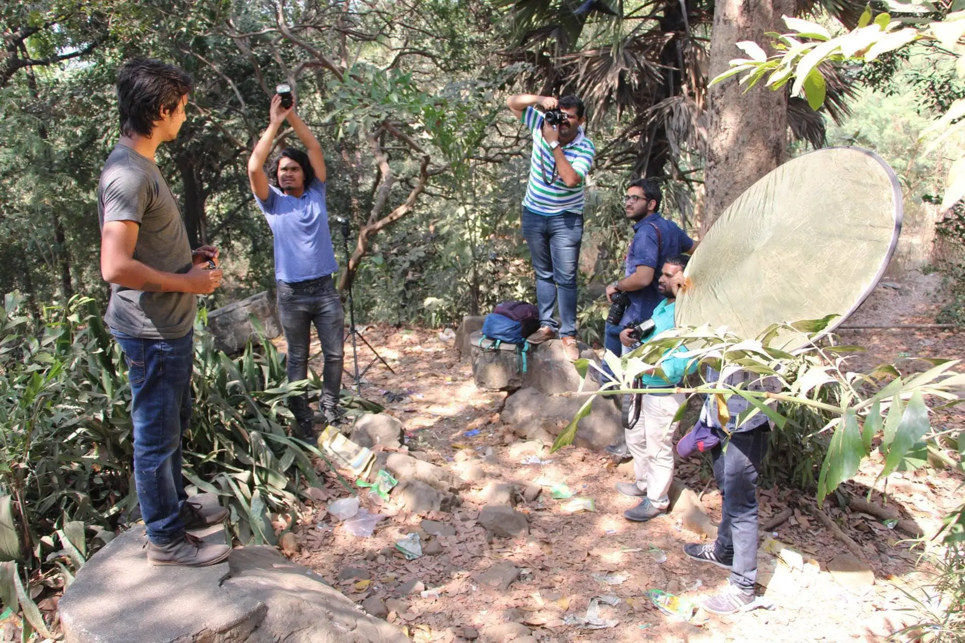 A group of individuals engaged in a photography session outdoors, surrounded by greenery and equipped with cameras and a reflector.