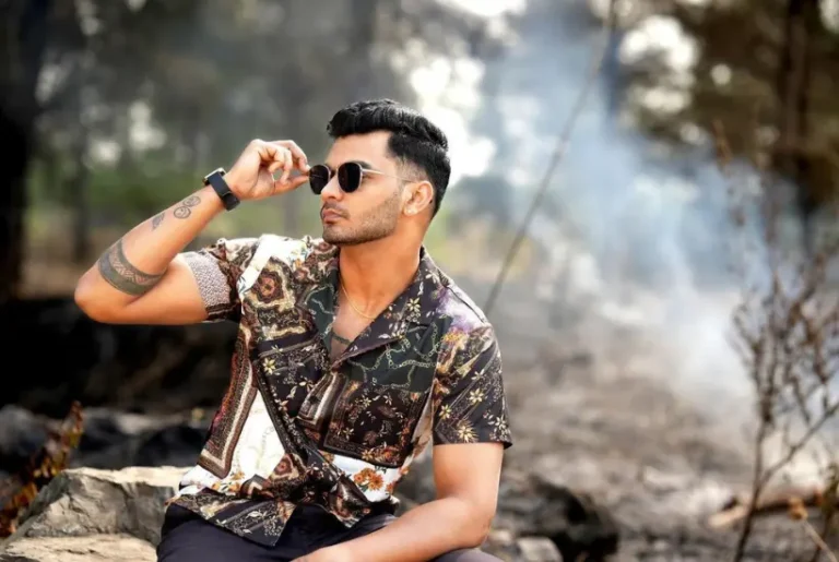 A man wearing sunglasses and a patterned shirt, sitting on a rock with a smoky background.