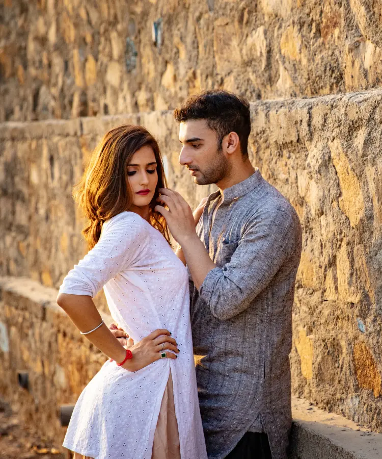 A couple posing against a stone wall, with the woman in a white outfit and the man in a traditional kurta.