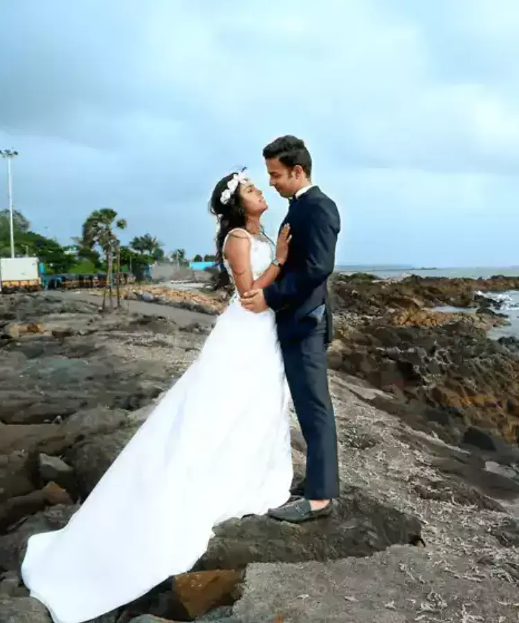 A couple in wedding attire standing on rocky terrain by the sea.