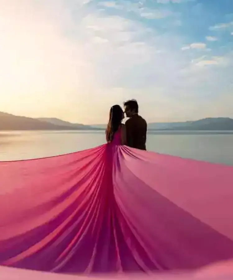 A couple standing by the water, with the woman wearing a flowing pink dress that extends behind her.