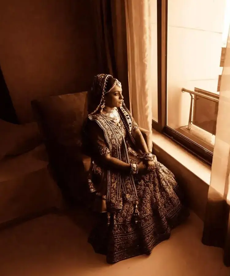 A sepia-toned photograph of a indian bride in traditional attire sitting beside a window, with their face obscured by a privacy bar.