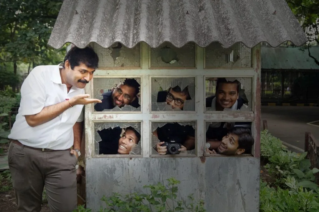 group of five people peeking through the windows of a rustic structure, with one man outside playfully gesturing towards them.