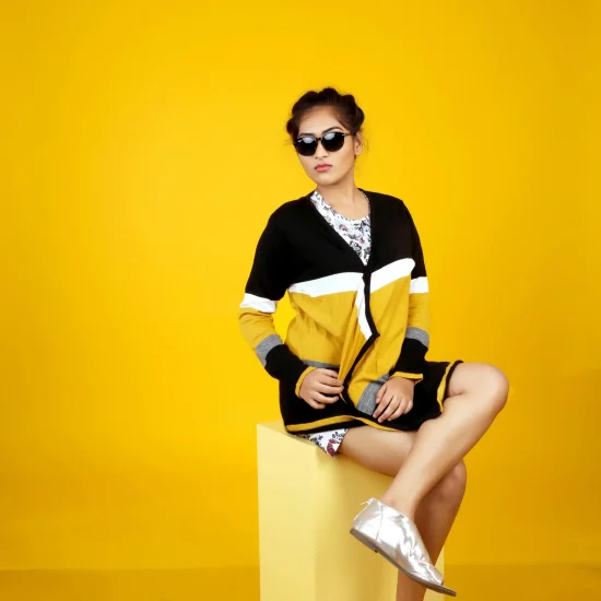 A person sitting on a yellow cube against a yellow background, wearing a black and white outfit with yellow accents and silver shoes.